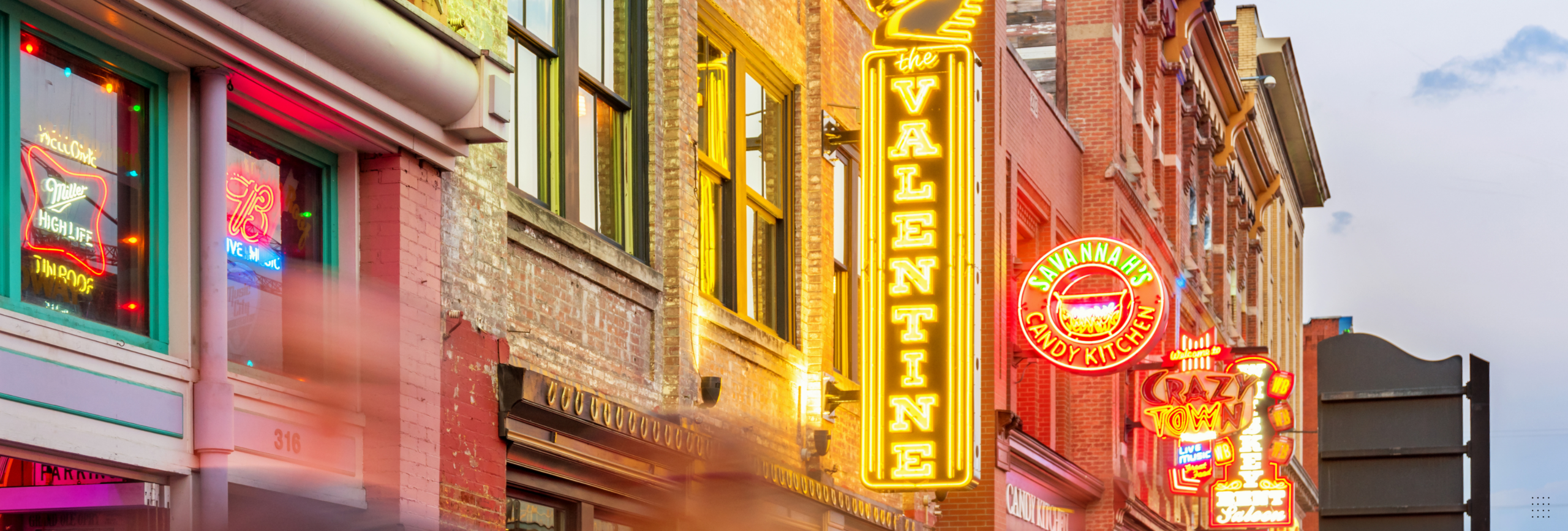 A vibrant street scene with bright neon signs illuminates the brick buildings, including Valentine and Nashville Crossroads. The lively atmosphere, enhanced by the blurred motion of pedestrians, reflects the energy of a community aware of mental health rehabs in Nashville.