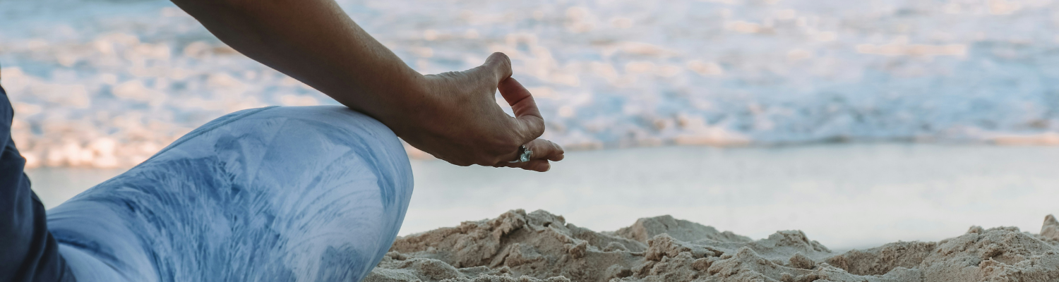 A person sits cross-legged on the sand, with the ocean whispering in the background. Their hand rests on their knee in a meditation pose, embodying a serene hypnotherapy session. The blue and white patterned leggings mirror the tranquil scene.