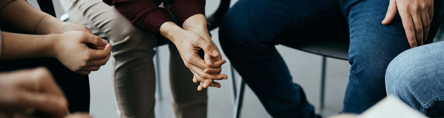 A group of people sit closely in a circle, only their hands and legs visible. Some hands are clasped, suggesting a supportive or thoughtful setting. The informal scene hints at Atlanta Mental Health Treatment Programs, possibly a meeting or group therapy session.