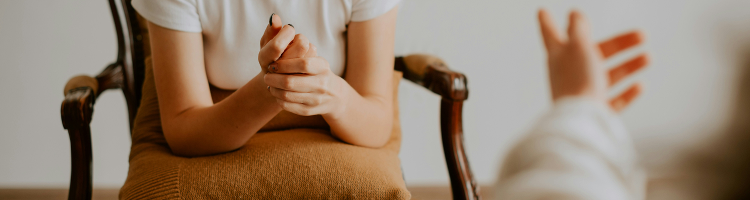 A person sits on a chair, resting their arms on a cushion, hands clasped. Another persons hand reaches toward the seated individual, evoking the supportive environment of Atlantas mental health treatment programs, suggesting a conversation or counseling session.