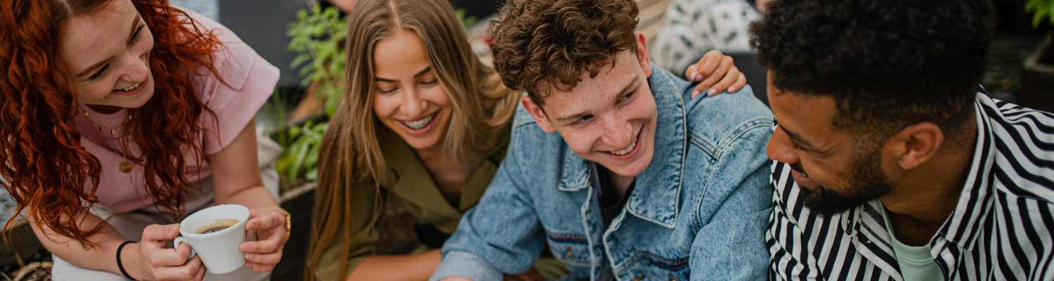 Four people sitting together outdoors, smiling and chatting. One person holding a cup of coffee. They appear relaxed and joyful, dressed in casual clothing, surrounded by greenery—a serene moment that brilliantly underscores the importance of mental health treatment for executives.