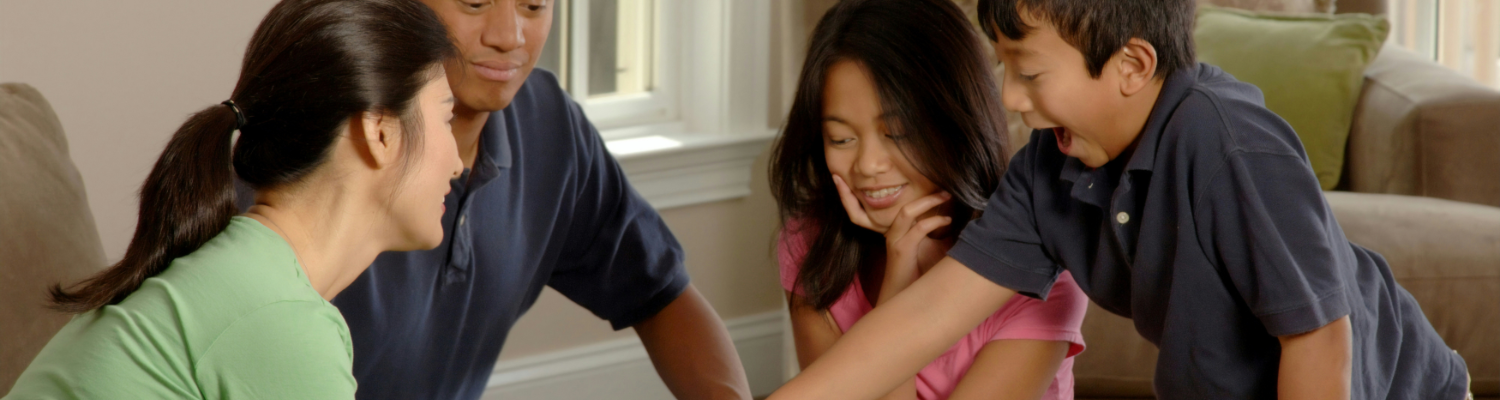 A family of four, including two adults and two children, is smiling and engaged in a board game at home. With the room well-lit by a window, they appear to be enjoying their time together—a moment of relaxation that even busy executives could appreciate for mental health.