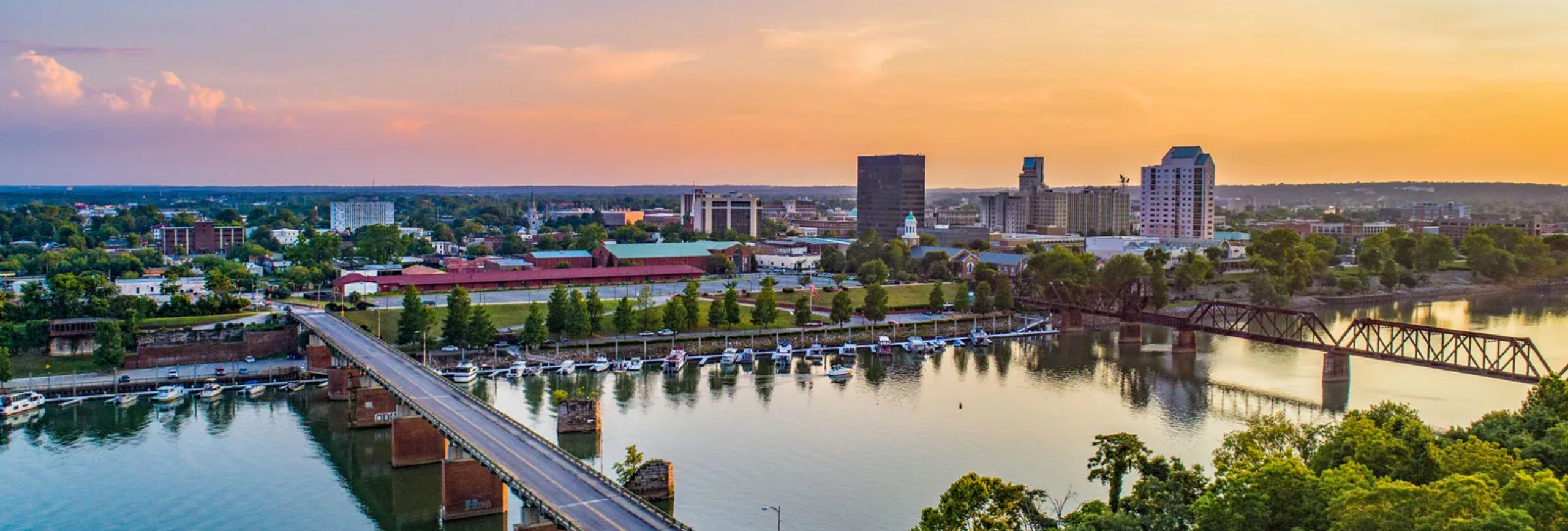 Aerial view of a cityscape at sunset, with buildings and a river where boats line the shore. The sky, like a canvas of warm hues, reflects on the water near trees bordering the banks. Its a serene scene perfect for reflection; even Jasper would find it therapeutic amidst his mental health treatment journey.