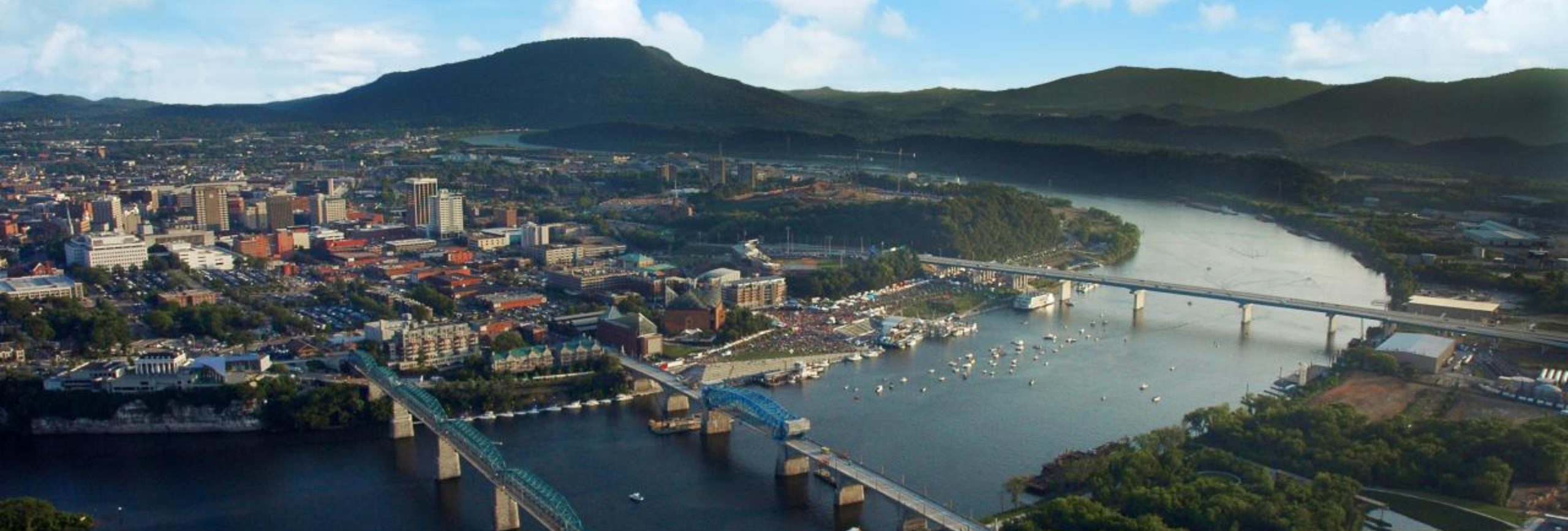 Aerial view of a city with multiple bridges crossing a river, surrounded by green areas and mountains in the background. The scene, reminiscent of the calm found in mental health treatment, is dotted with small boats under a clear sky that enhances the scenic landscape.