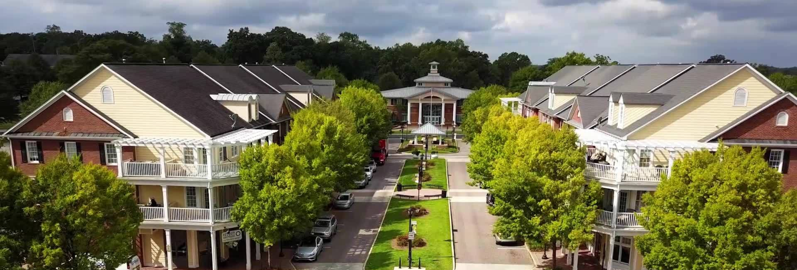Aerial view of a tree-lined street flanked by two buildings with beige siding and brick accents, each offering mental health treatment in Marrietta. Both have multiple floors and balconies. A circular building with a tower is visible at the streets end, set against a backdrop of dense trees.