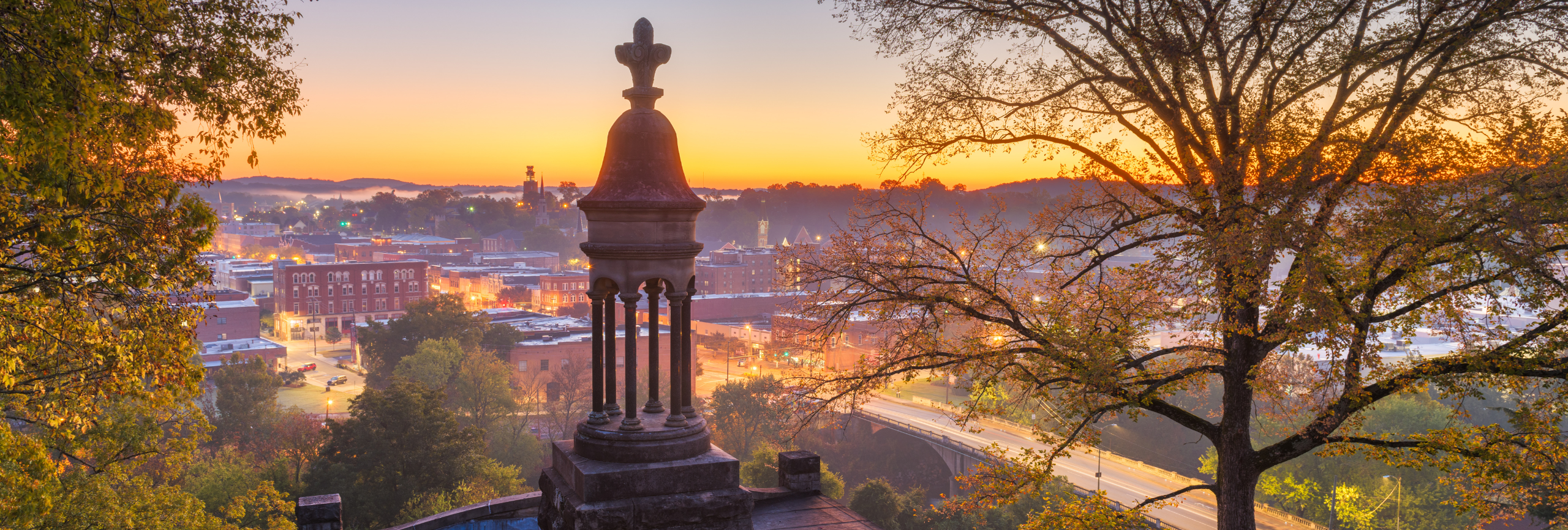 A scenic sunrise view over a small town with historic brick buildings, a stone monument in the foreground, trees framing the image, and a soft orange sky in the background—capturing the peaceful spirit of Mental Health Treatment in Rome.