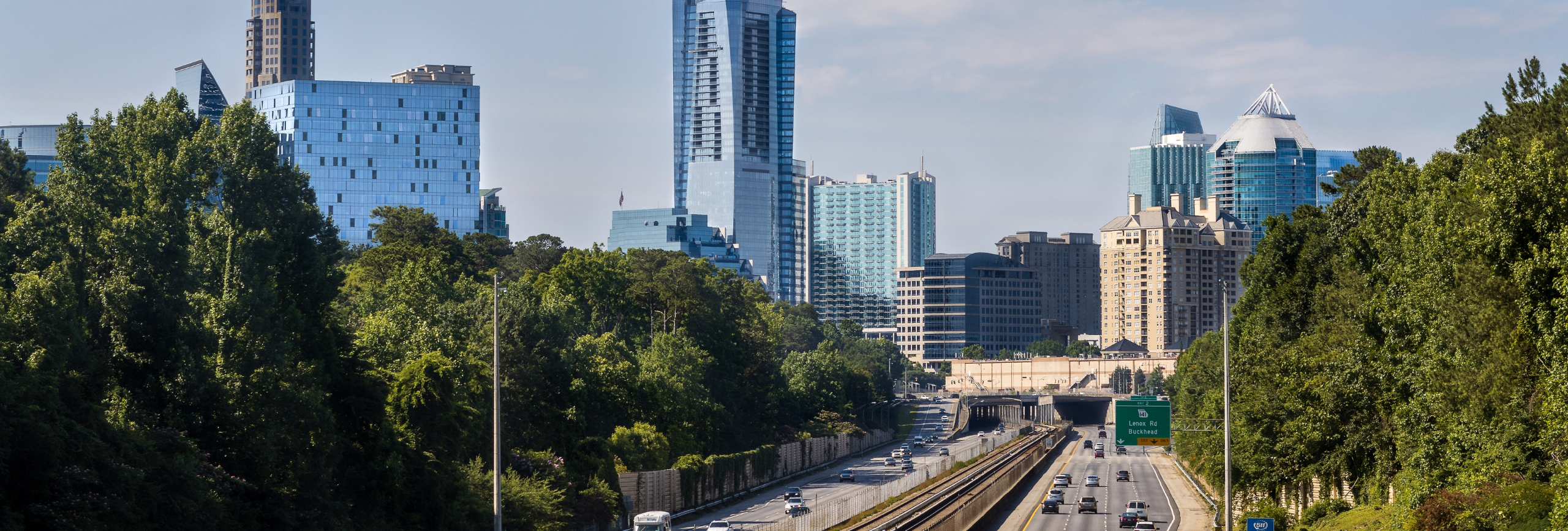 A city skyline with tall modern buildings rises behind a freeway in Buckhead, where cars travel in both directions, flanked by green trees under a clear blue sky—a vibrant setting for mental health treatment in Buckhead.